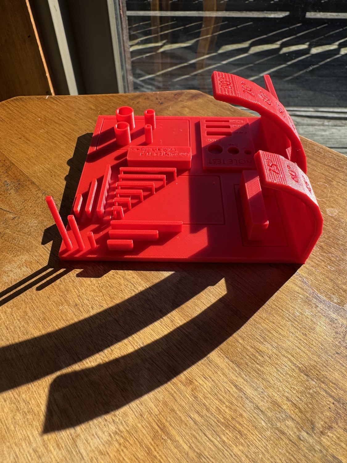 Red 3D printer test with overhangs, fine walls, and cylinders on a sunlit wooden table